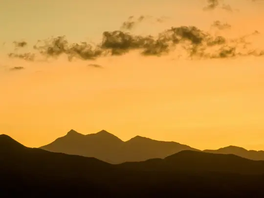 A landscape photo showing dark mountain silhouettes against a bright orange and yellow sky with dark, wispy clouds.