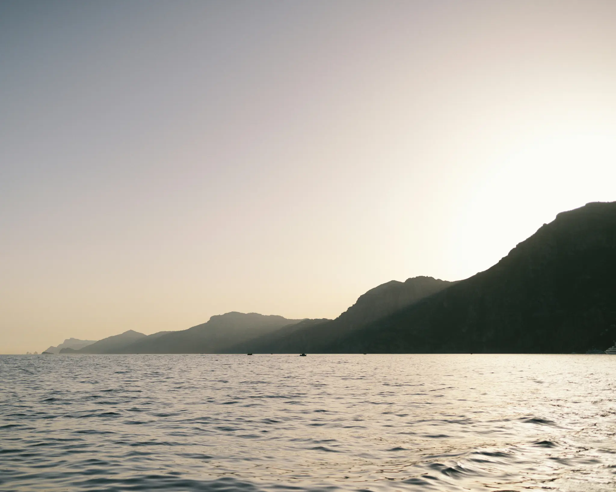 A wide landscape photo showing a calm sea in the foreground, with a range of dark mountains silhouetted against a bright…