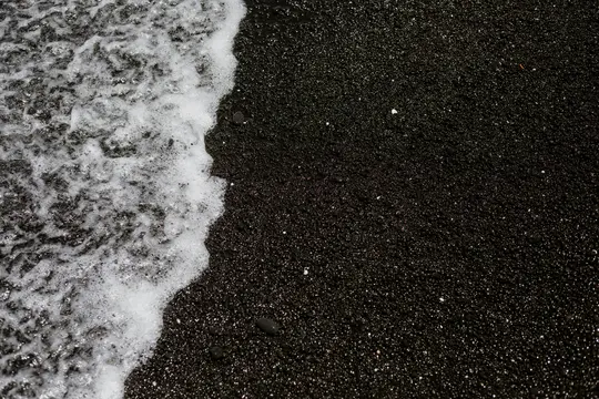 Vista ravvicinata, dall'alto, di un'onda bianca e schiumosa che si ritira da una spiaggia scura di sabbia nera, creando un