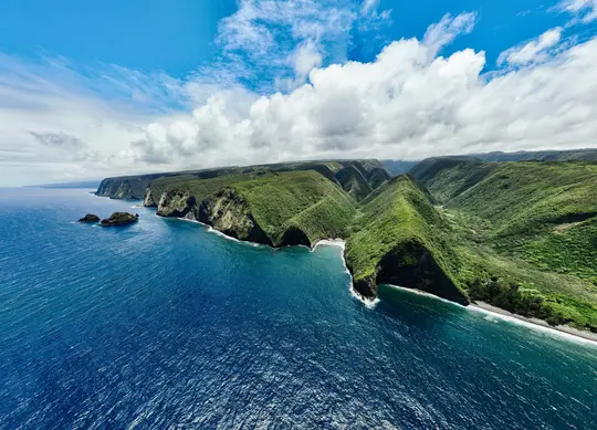 An aerial photograph captures a dramatic coastline where verdant, steep cliffs and valleys meet the deep blue ocean. White…