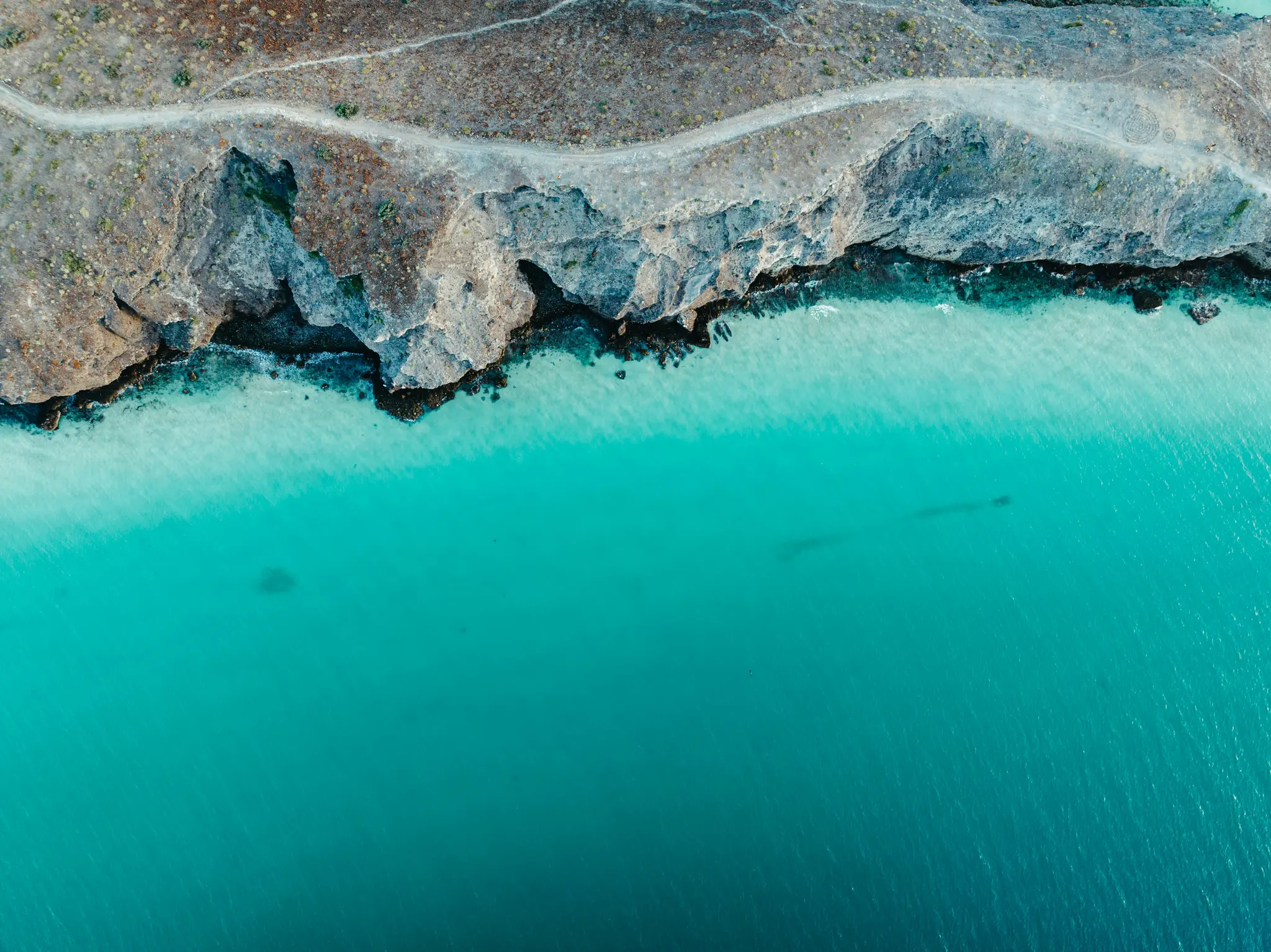 En flygfotografi visar en karg kustlinje med grå klippor och en grusväg ovanpå, som möter klart turkost havsvatten.