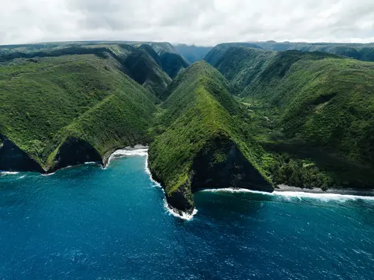 An aerial view shows a deep blue ocean meeting a coastline of steep, verdant green valleys and dark sand beaches under a…