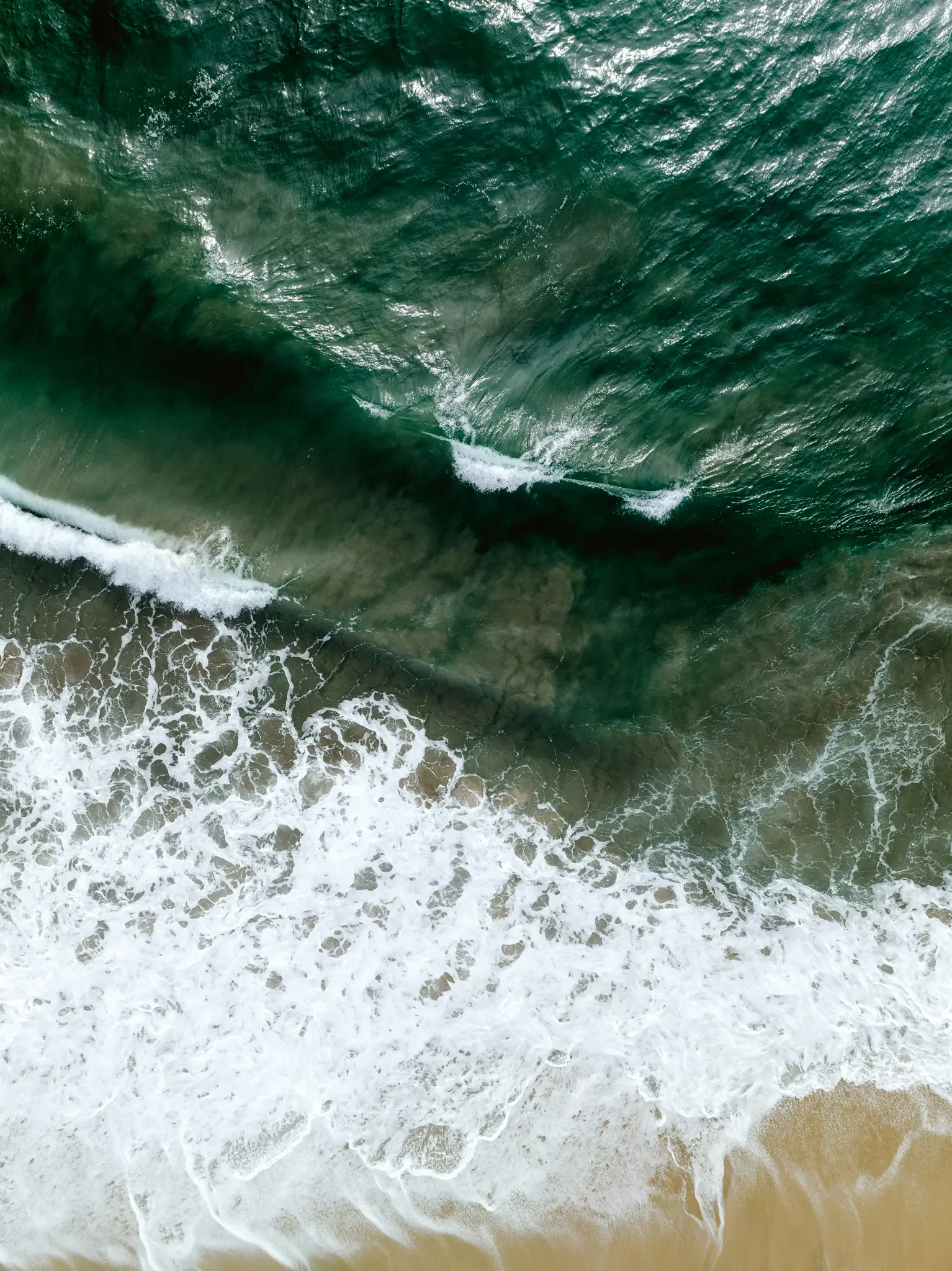 En flygbild som ser ner på havet och visar djupt gröna vågor som bryts till vitt skum på en ljusbrun sandstrand.