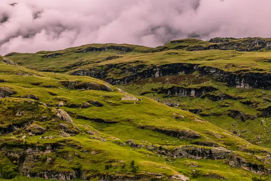 Breite Landschaftsaufnahme von üppig grünen Hügeln mit sichtbaren Felsvorsprüngen und Felsbändern unter einem Himmel voller