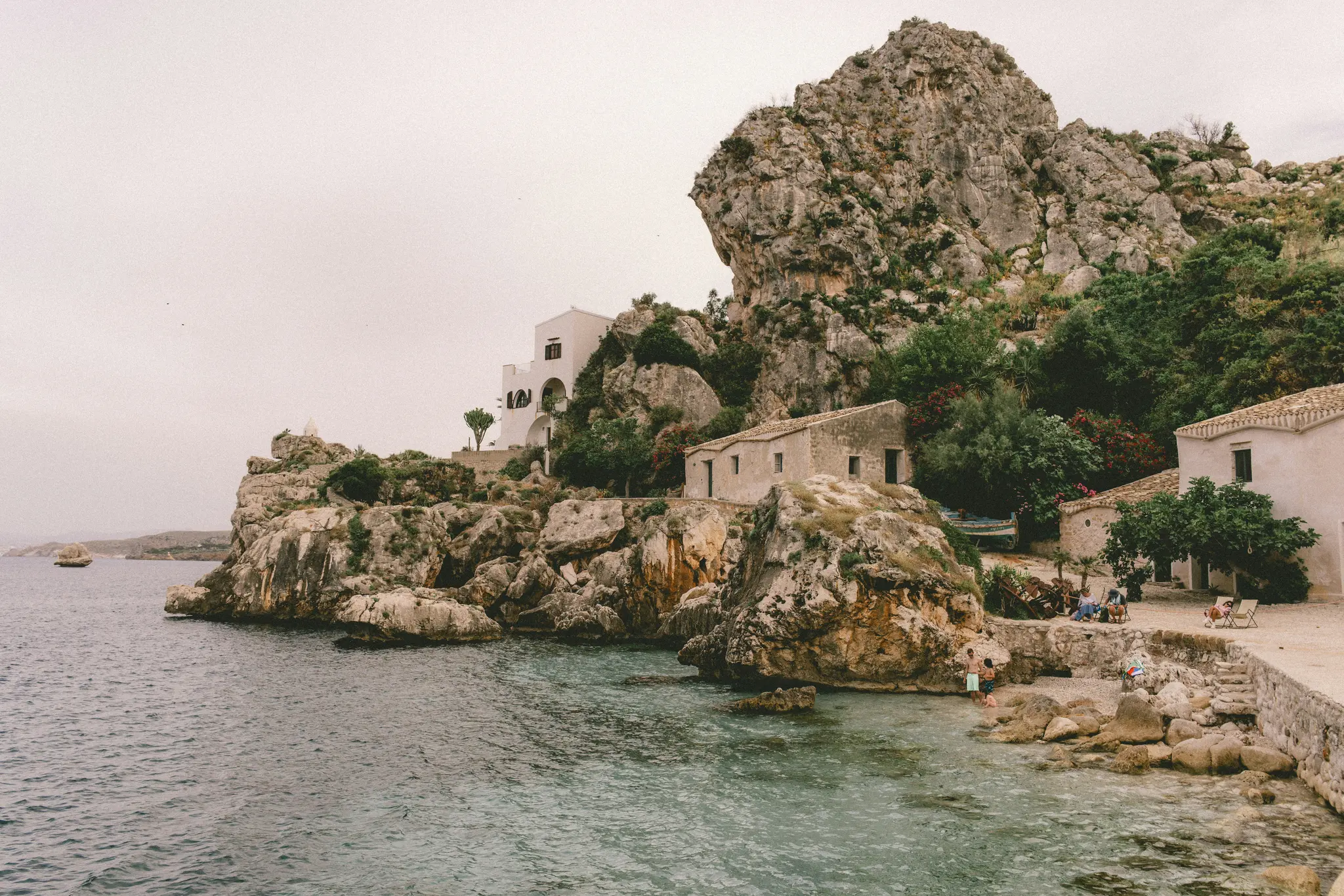 A wide landscape photo of a rocky coastline with several stone and white buildings, lush green vegetation, and a small…