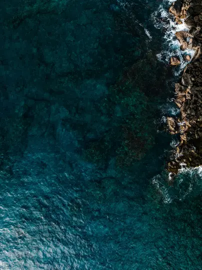 An aerial, top-down view shows deep blue and teal ocean water meeting a rugged, dark brown rocky coastline. White waves…