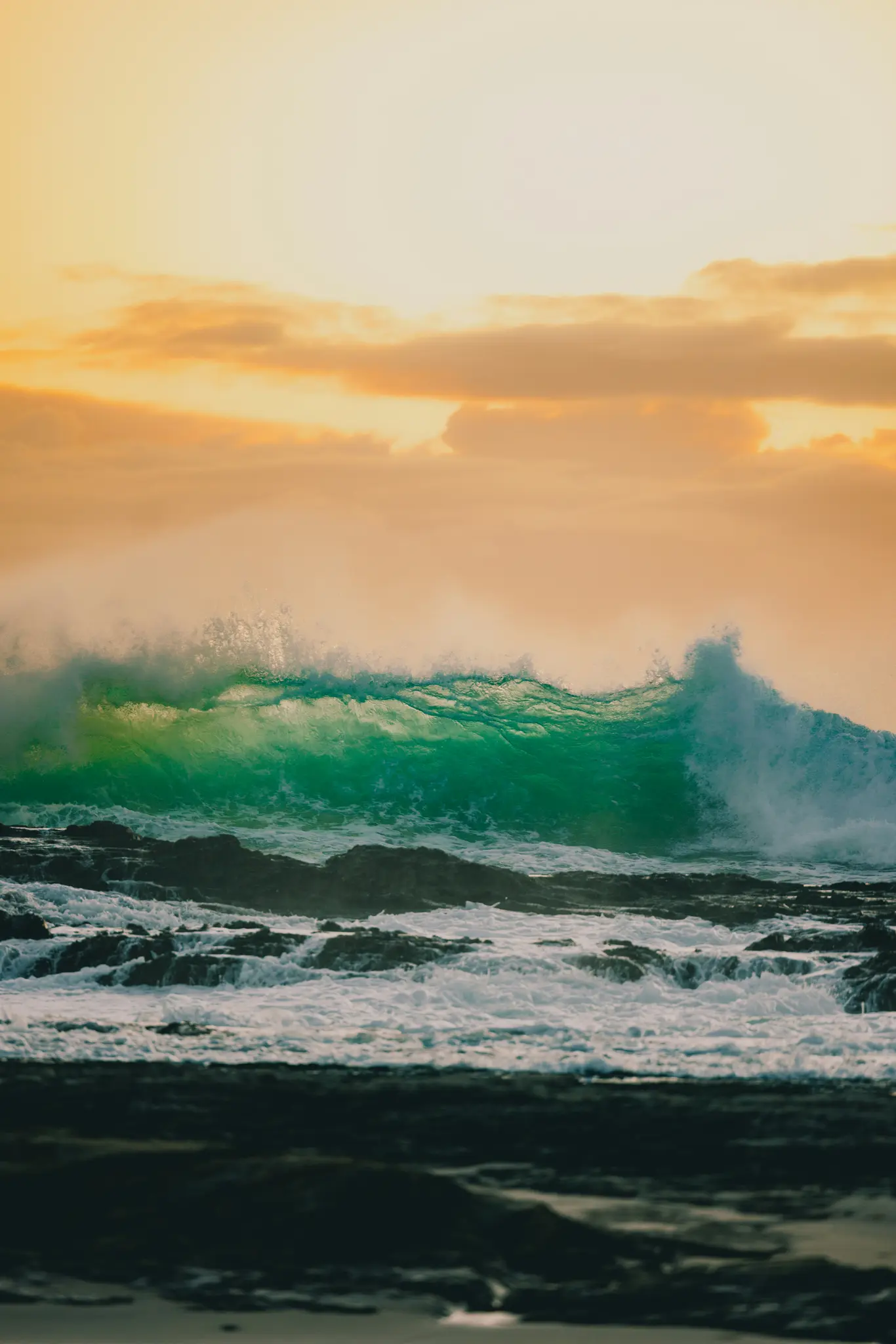 A large, translucent green wave crests and crashes over dark, jagged rocks, with white foam at its base. The sky above is a…