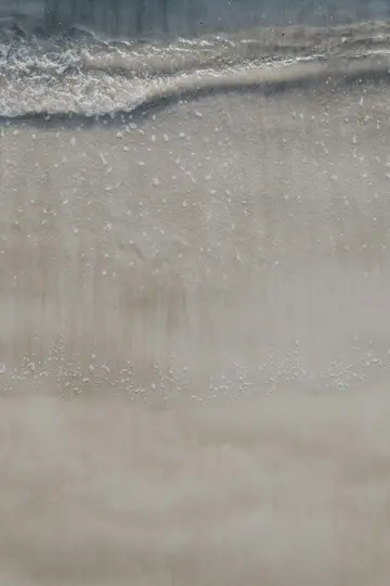 Eine Nahaufnahme von oben eines Sandstrandes mit einer kleinen, zurückweichenden Welle, die eine Linie aus weißem Schaum und