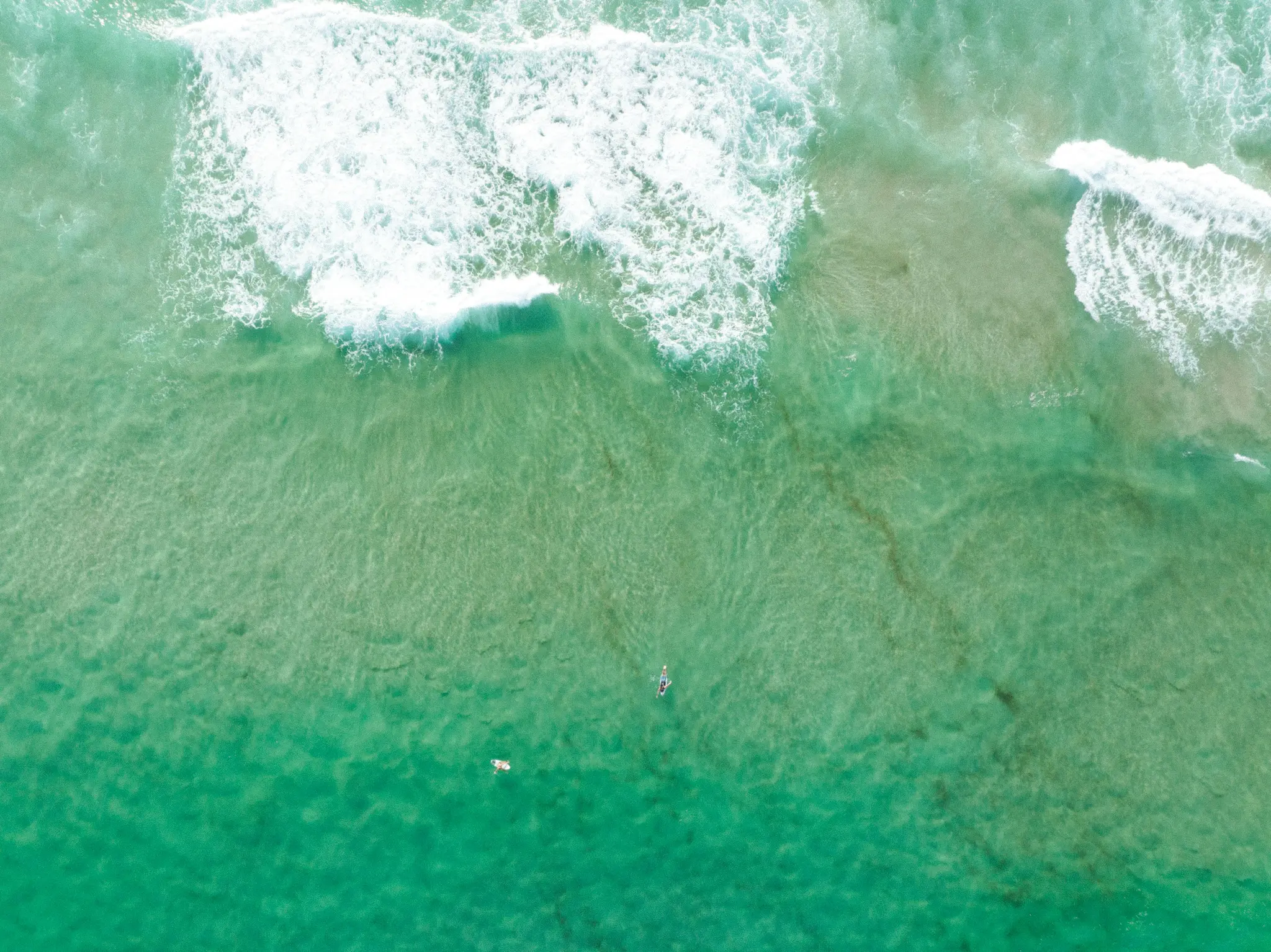 Snimka iz zraka na bistri tirkizni ocean s bijelim valovima koji se lome. Dva mala paddleboardera vidljiva su u mirnoj vodi.
