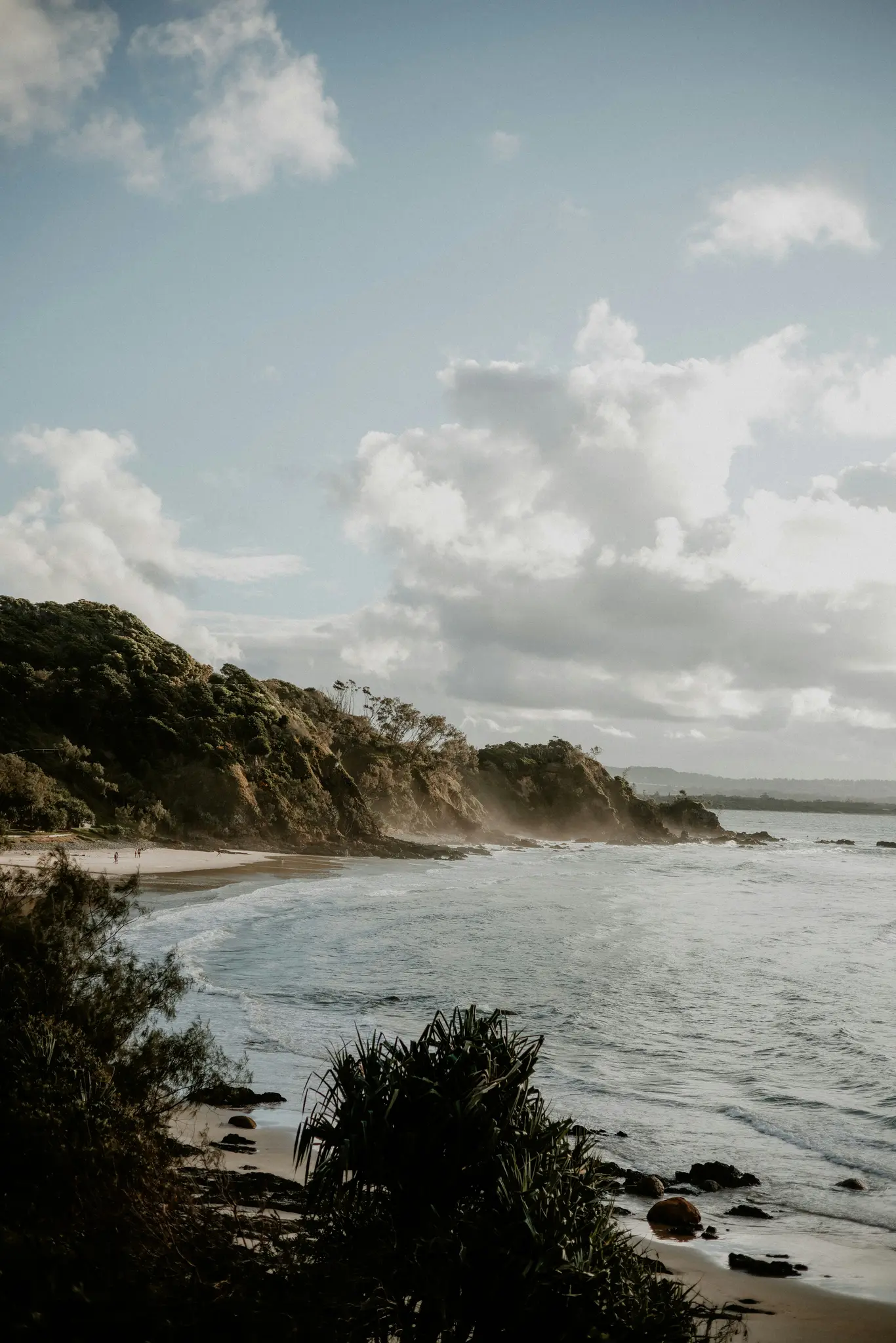 A wide landscape photo of a beach with gentle waves, a steep green cliff, and a cloudy sky. Three small figures are on the…