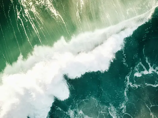 Uno scatto aereo di una potente onda oceanica che si infrange. Creste bianche e spumose si estendono diagonalmente su acqua