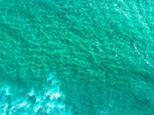 Eine Luftaufnahme zeigt klares türkisfarbenes Meerwasser mit Wellen und Mustern auf der Oberfläche und sichtbarem Sandboden