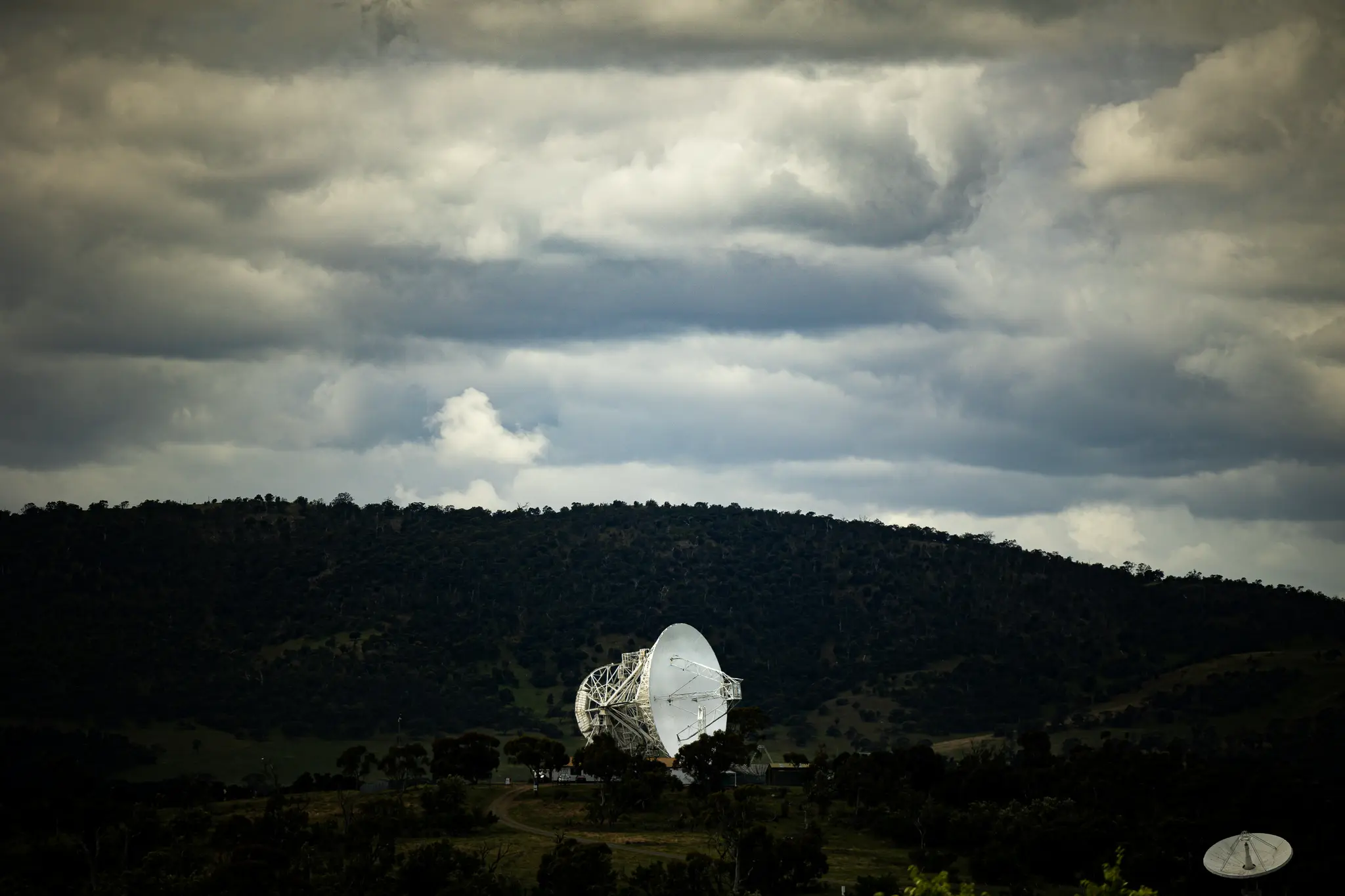 A large white radio telescope dish stands prominently in a valley, surrounded by dark green, tree-covered hills under a…