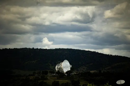 A large white radio telescope dish stands prominently in a valley, surrounded by dark green, tree-covered hills under a…