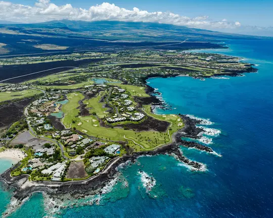 An aerial photo shows a lush green golf course and white houses nestled among dark volcanic rock along a vibrant blue…