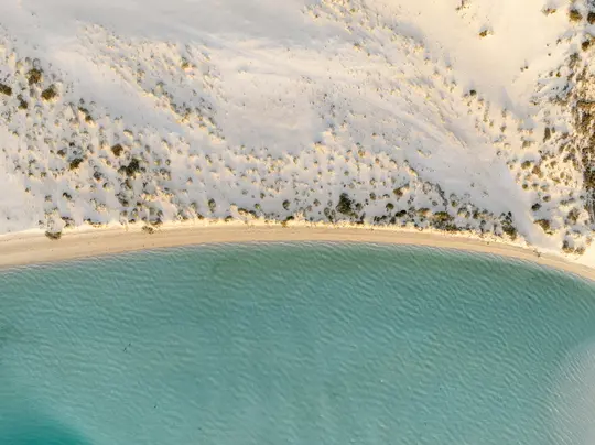Eine Luftaufnahme zeigt einen geschwungenen Sandstrand mit spärlicher grüner Vegetation, der auf ruhiges, klares