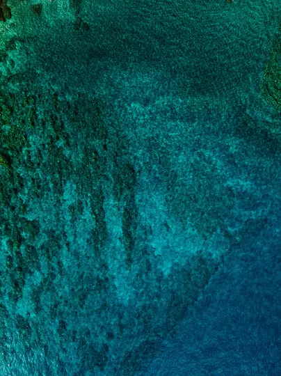 Una fotografia aerea mostra acqua oceanica limpida con una sfumatura di tonalità blu e verdi, che rivela il fondale marino