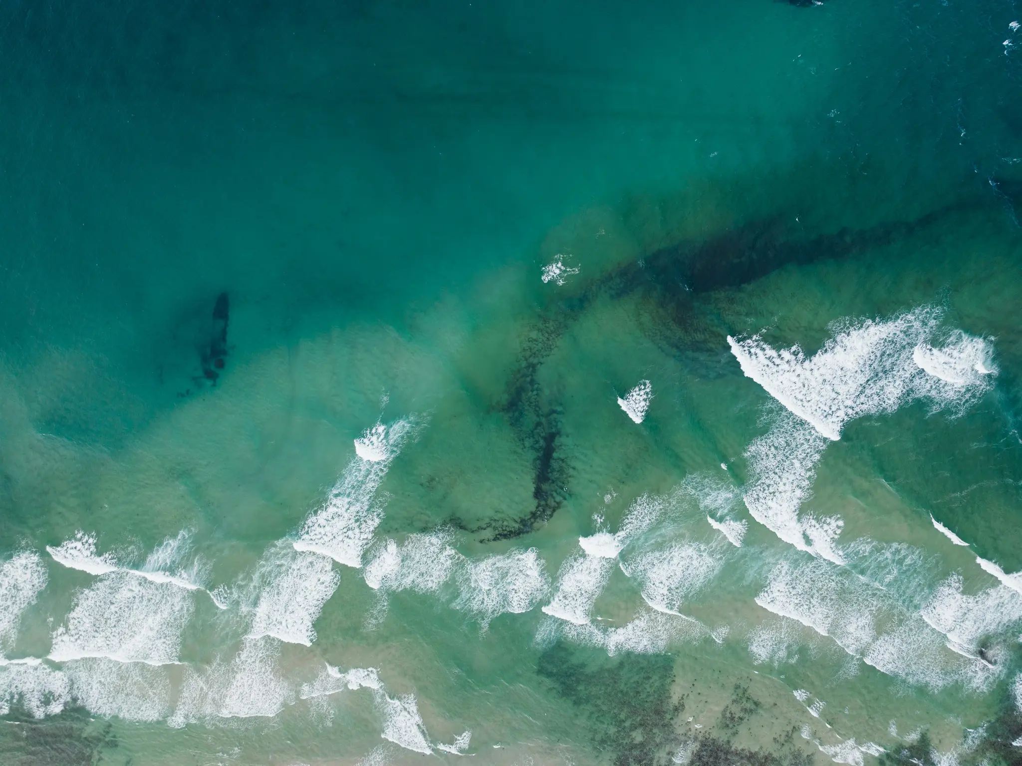 En top-down luftudsigt over havet, der viser bølger, der bryder i hvidt skum over en lavvandet, grønblå havbund.