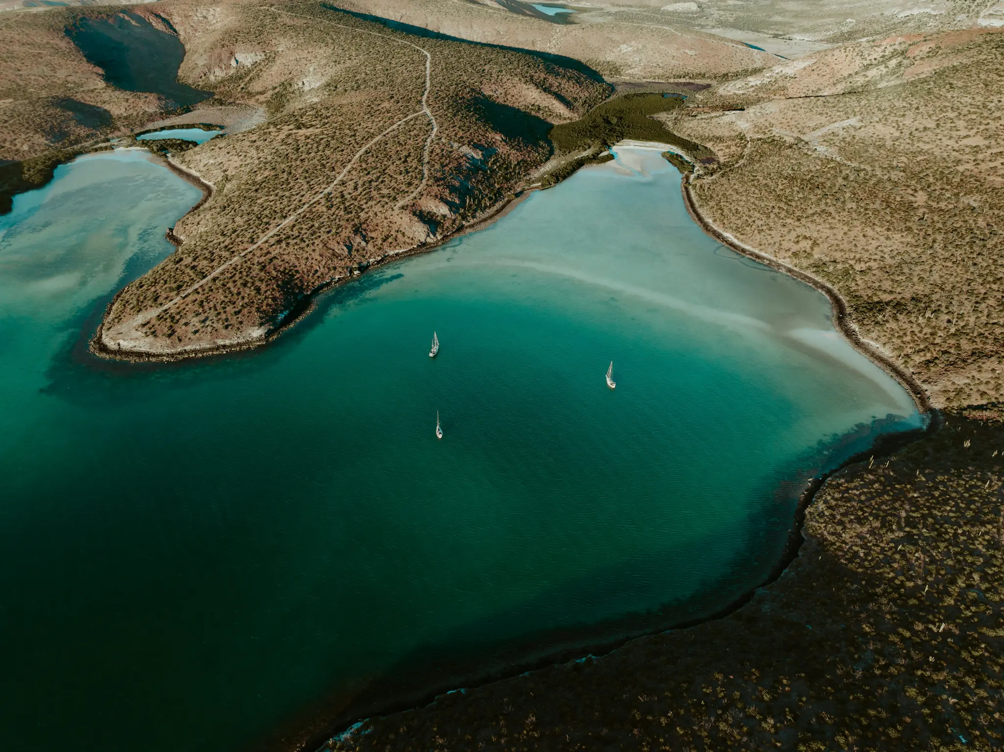 An aerial shot captures three small white sailboats on a calm, deep turquoise bay, bordered by dry, brush-covered hills.