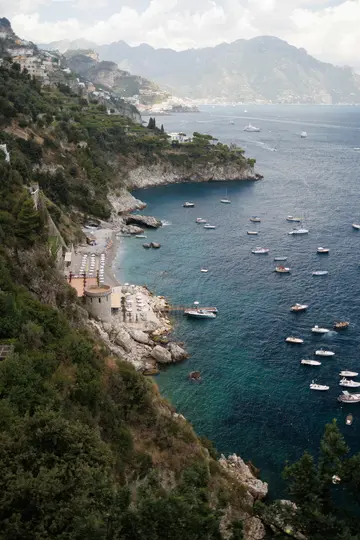 An aerial view shows a vibrant blue bay dotted with numerous boats, bordered by a steep, green hillside with a town.