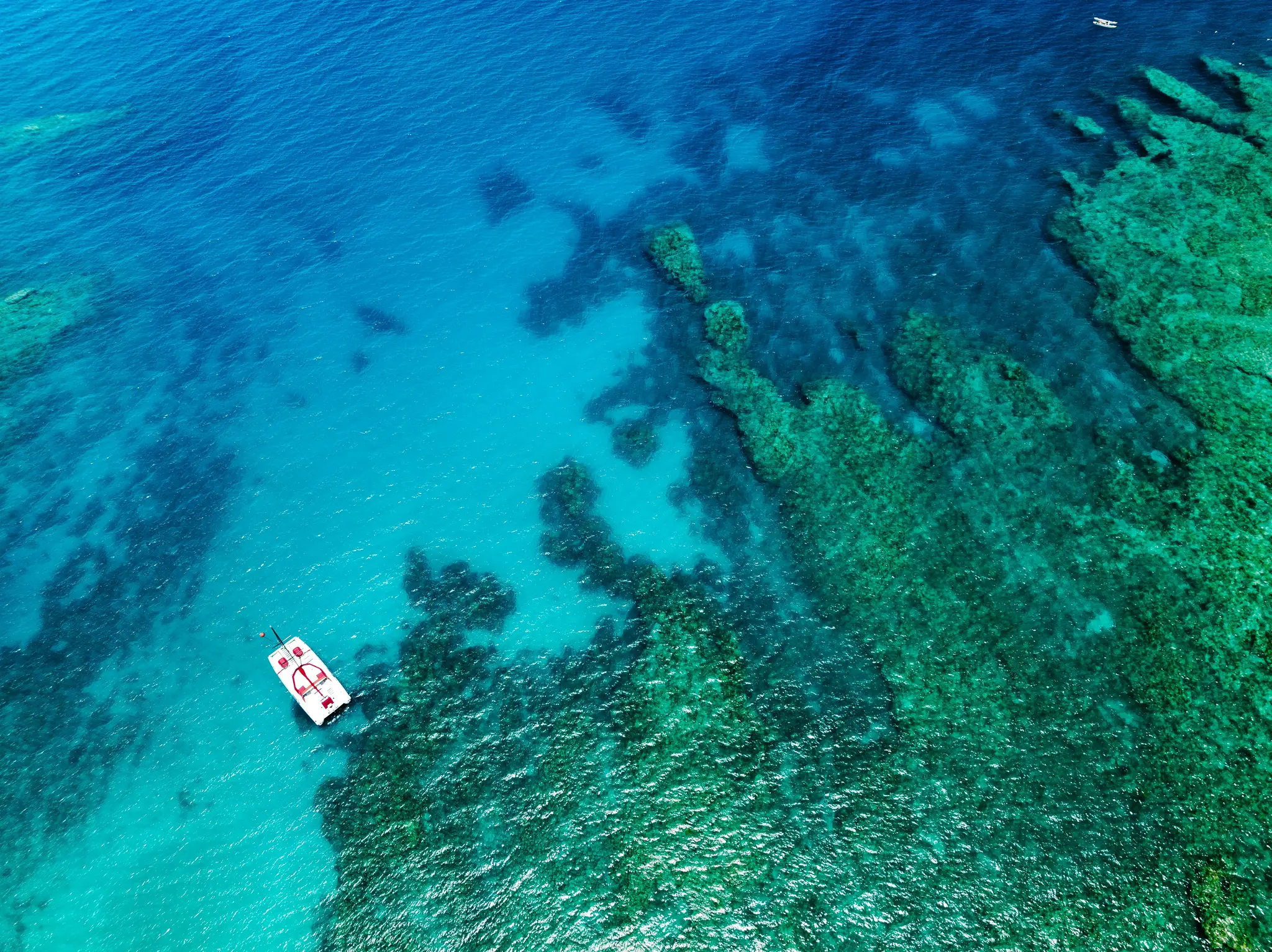 An aerial shot captures a white catamaran with red accents floating on clear turquoise water next to a vibrant green coral…
