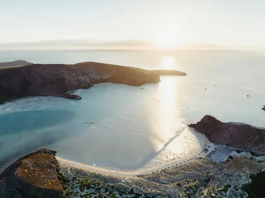 An aerial photo shows a calm bay with clear turquoise water reflecting the golden light of the setting sun. Arid hills…
