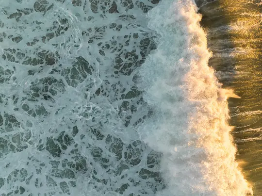 Un'onda bianca e frangente nell'oceano, illuminata da una luce dorata, vista dall'alto.