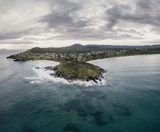An aerial view captures a coastal town with houses nestled among green trees, a long sandy beach, and a rocky headland.
