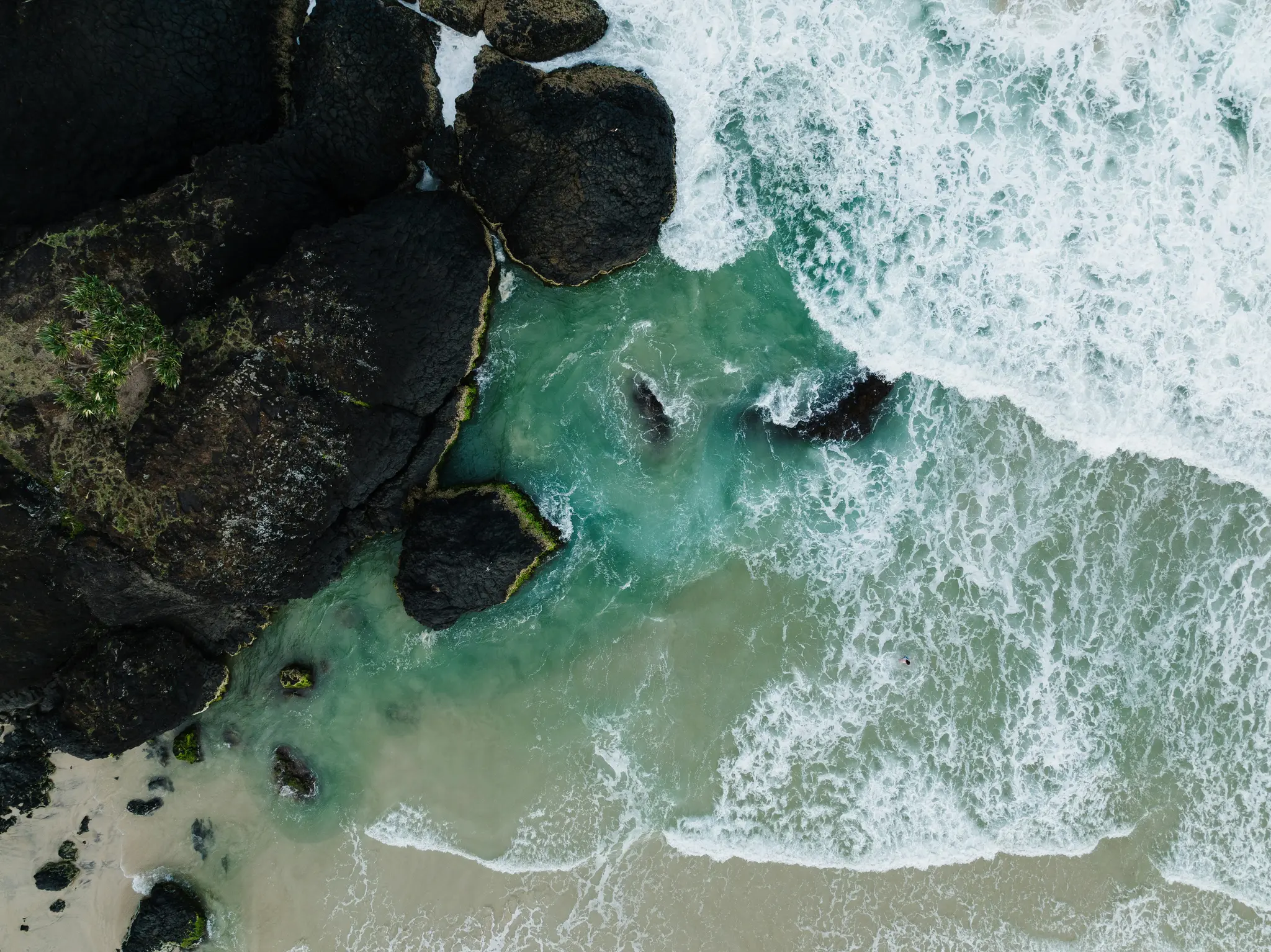 An aerial shot shows dark, textured rocks on the left, meeting turquoise and teal ocean water. White waves crash onto a…