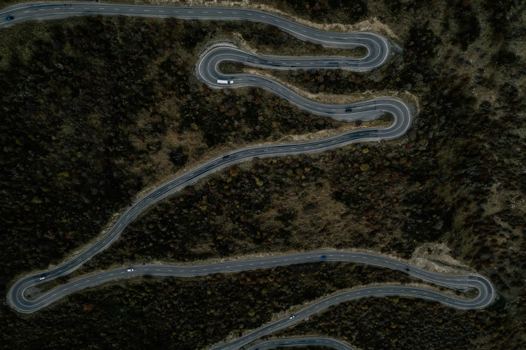 Een luchtfoto, van bovenaf, van een grijze asfaltweg met witte lijnen, die zich in meerdere haarspeldbochten door een