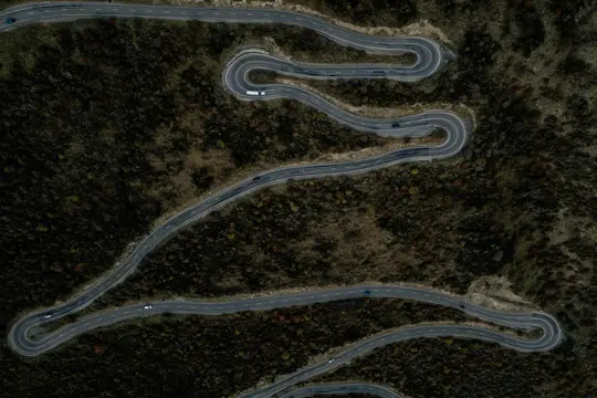 Una vista aerea, dall'alto, di una strada asfaltata grigia con linee bianche, che si snoda in molte curve a tornante