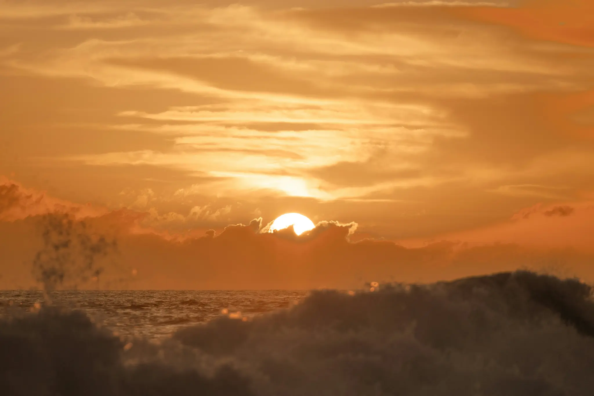 Ein lebendiger Sonnenuntergang über dem Ozean, die Sonne teilweise von Wolken verdeckt. Im Vordergrund sind große Wellen zu