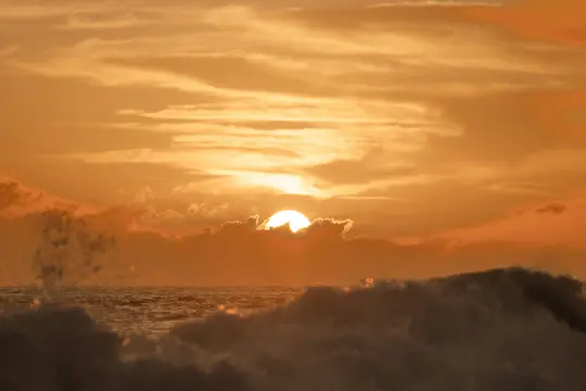 Un vibrante tramonto sull'oceano, il sole parzialmente nascosto dalle nuvole. Grandi onde sono visibili in primo piano.