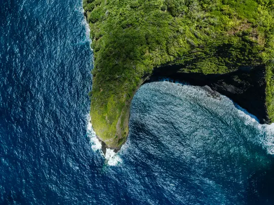 An aerial view shows a vibrant green, tree-covered peninsula jutting into a deep blue ocean. Waves crash against the rocky…