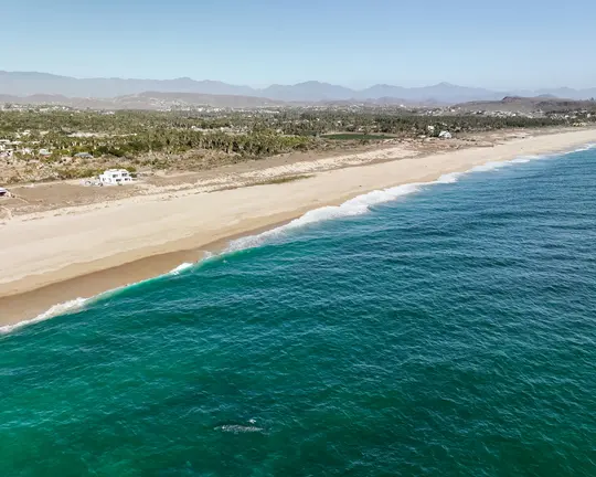 An aerial shot shows a long, light-colored sandy beach meeting vibrant turquoise ocean water with gentle waves. A coastal…