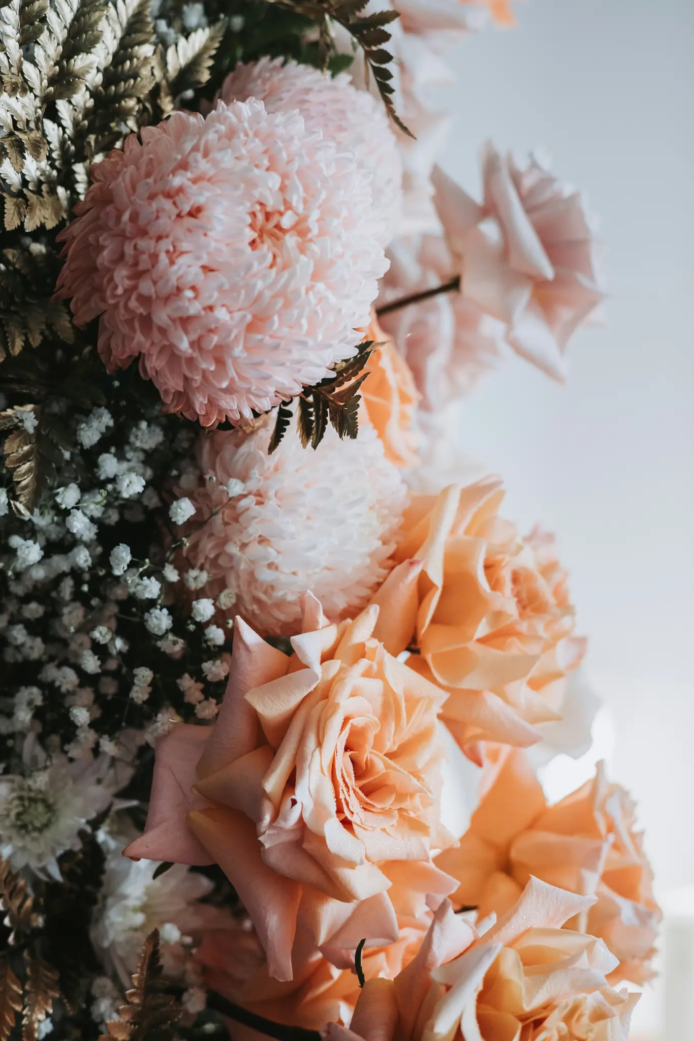 Et nærbillede af et blomsterarrangement med lyserøde krysantemum, ferskenfarvede roser, gypsophila og mørke bregner.