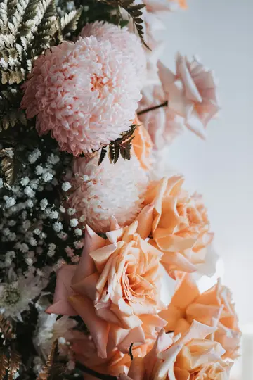 Eine Nahaufnahme einer Blumenanordnung mit hellrosa Chrysanthemen, pfirsichfarbenen Rosen, Schleierkraut und dunklen Farnen.