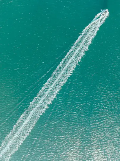 Vista aerea di una barca bianca che sfreccia su una vasta distesa di acqua turchese, lasciando una lunga scia bianca e