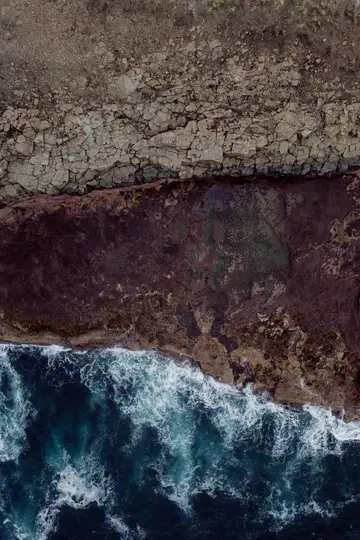 Una vista aerea, dall'alto, di onde oceaniche blu scuro con schiuma bianca che si infrangono su una costa rocciosa, aspra e