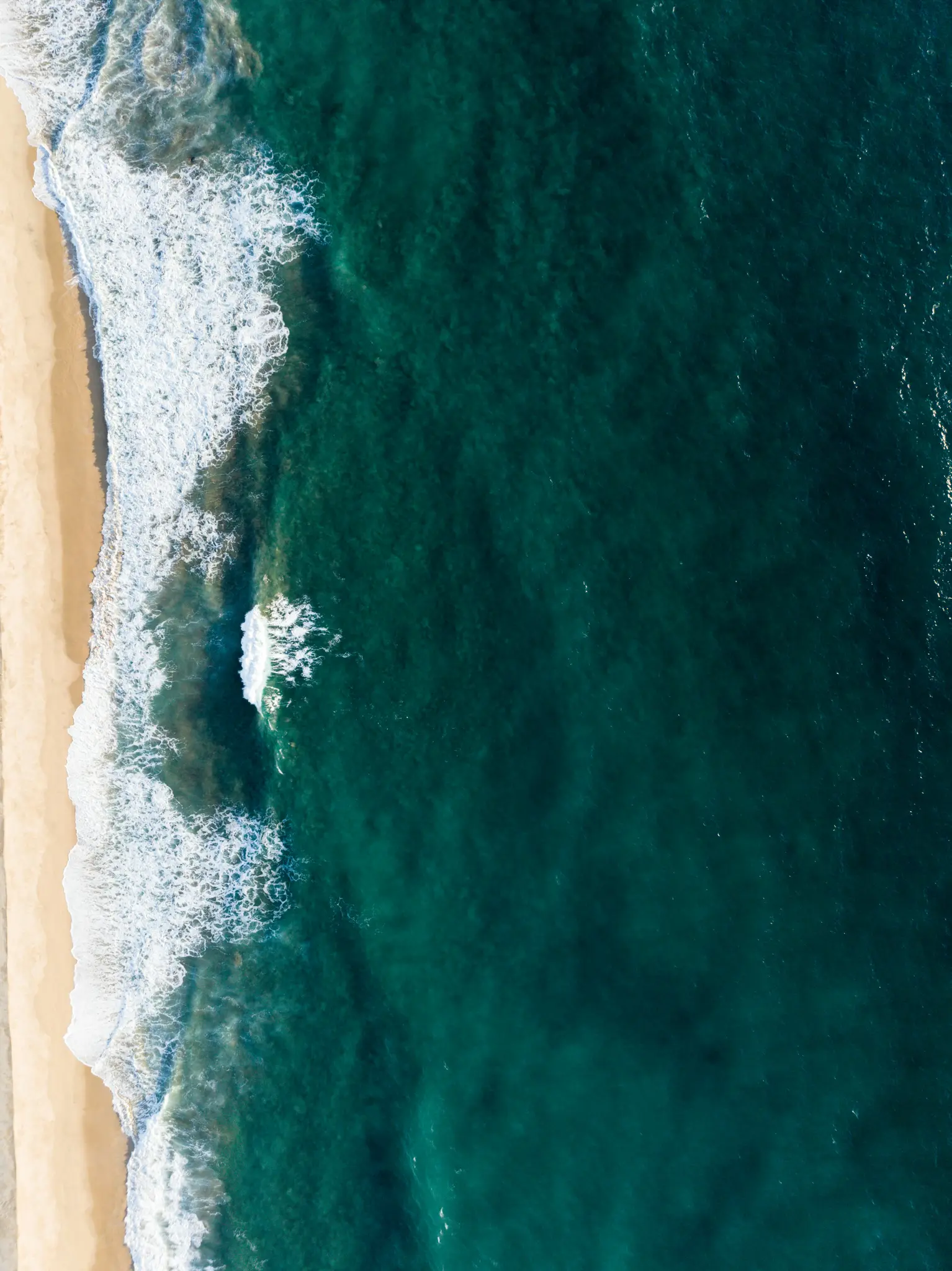 Una vista aérea, de arriba hacia abajo, muestra el océano azul verdoso profundo encontrándose con una playa de arena beige.