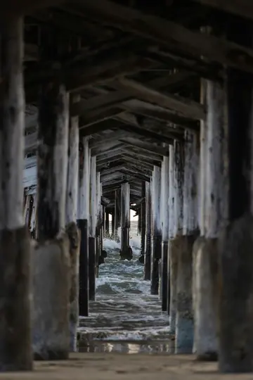 A low-angle view from under a weathered wooden pier, showing rows of dark pilings extending into the distance. Waves crash…