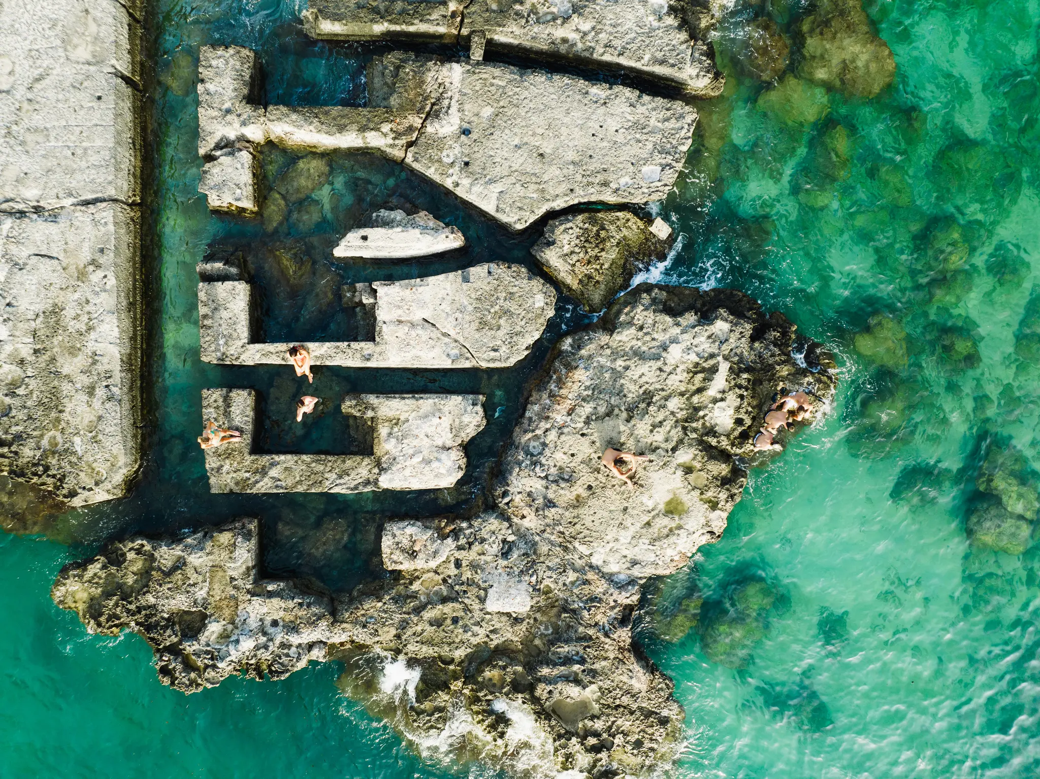 An aerial photo shows ancient-looking stone structures partially submerged in clear turquoise water, with six people…