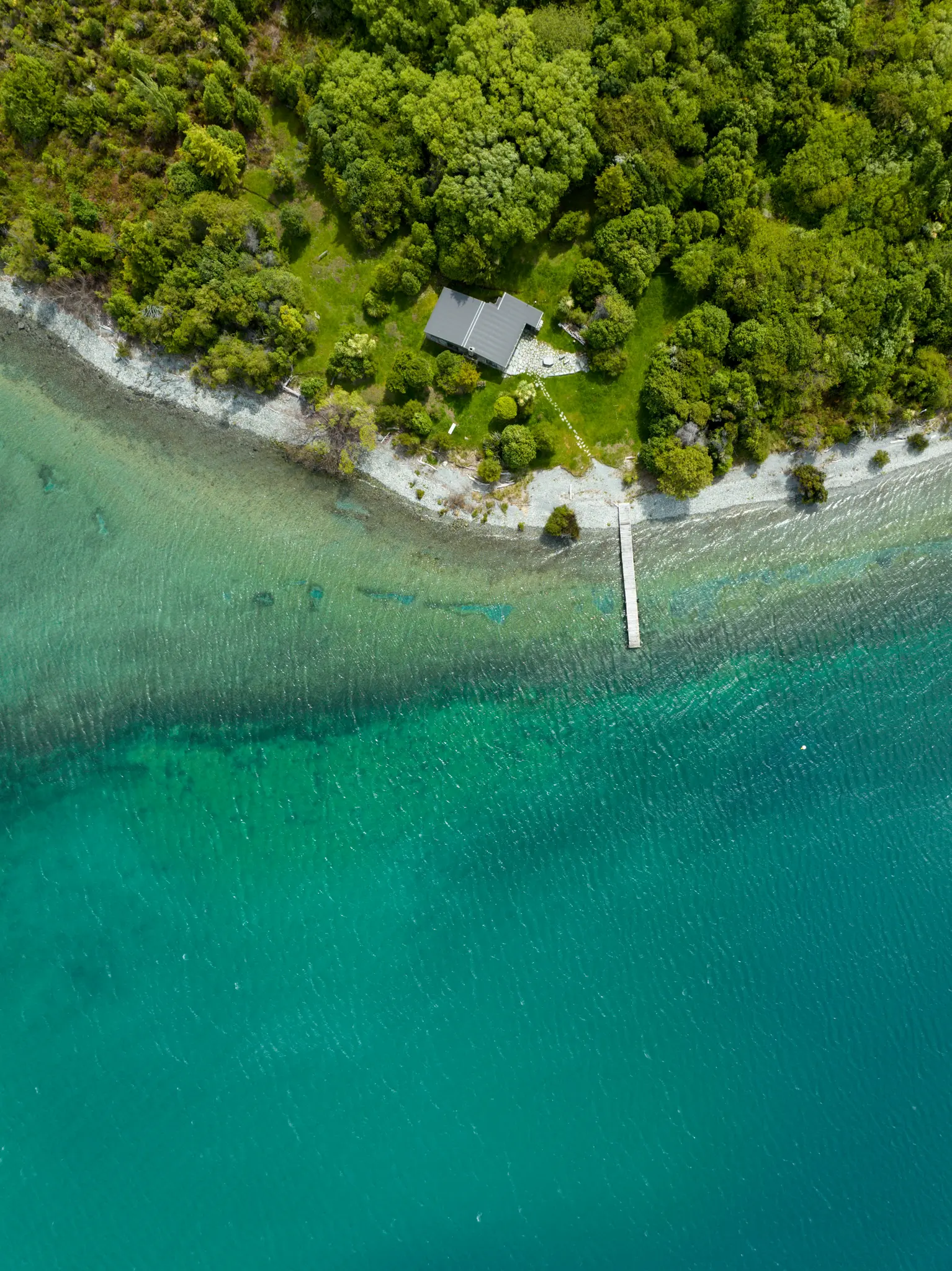 An aerial shot of a house with a gray roof surrounded by lush green trees on a rocky shore next to clear turquoise water…