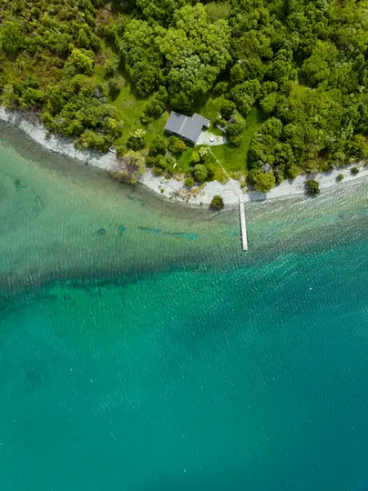 An aerial shot of a house with a gray roof surrounded by lush green trees on a rocky shore next to clear turquoise water…