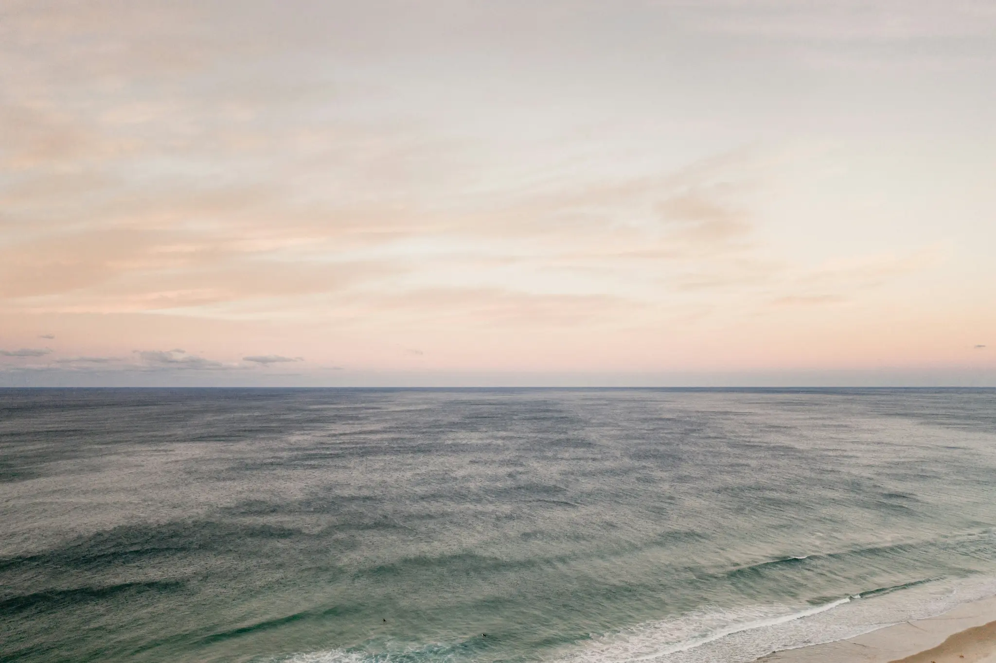 A wide landscape photo of the ocean under a soft, pastel-colored sky. Gentle waves roll towards a narrow strip of sandy…