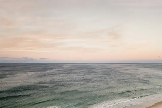 A wide landscape photo of the ocean under a soft, pastel-colored sky. Gentle waves roll towards a narrow strip of sandy…