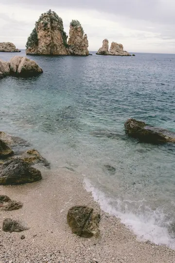 Un sereno paesaggio di una spiaggia di ciottoli con acqua limpida blu-verde che lambisce dolcemente la riva. Diverse grandi e