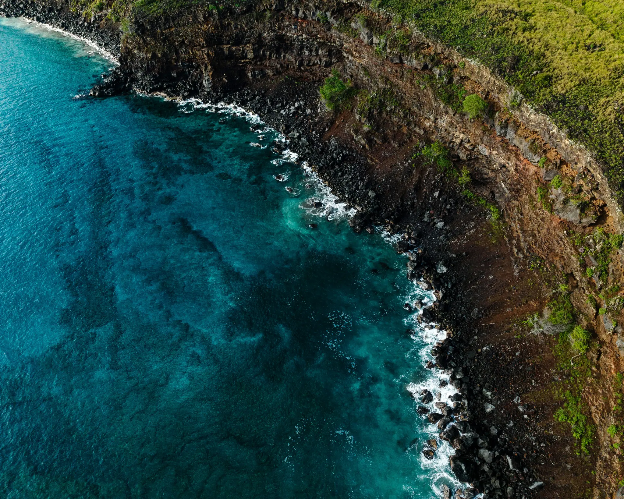 An aerial shot captures a dramatic coastline where dark, jagged rocks meet vibrant turquoise ocean water. Green vegetation…