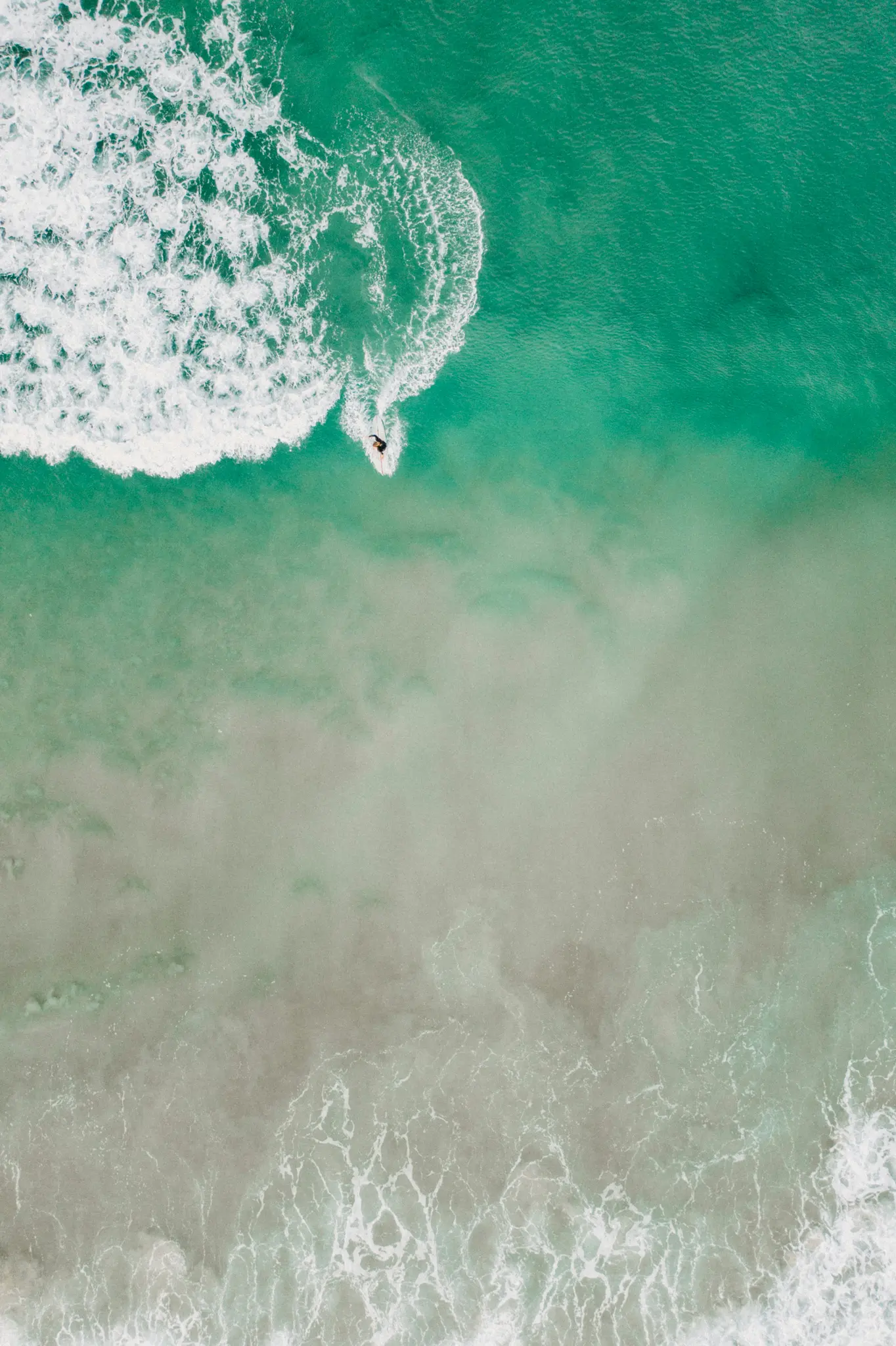 Eine Luftaufnahme zeigt einen einsamen Surfer auf einer weiß aufschäumenden Welle in lebhaftem türkisfarbenem und grünem