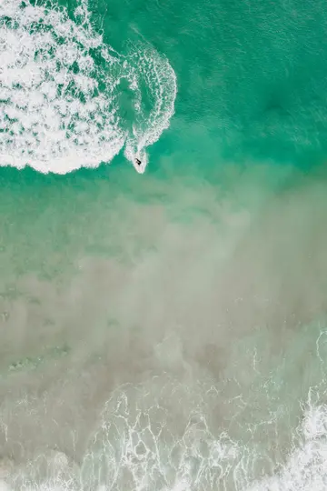 Eine Luftaufnahme zeigt einen einsamen Surfer auf einer weiß aufschäumenden Welle in lebhaftem türkisfarbenem und grünem